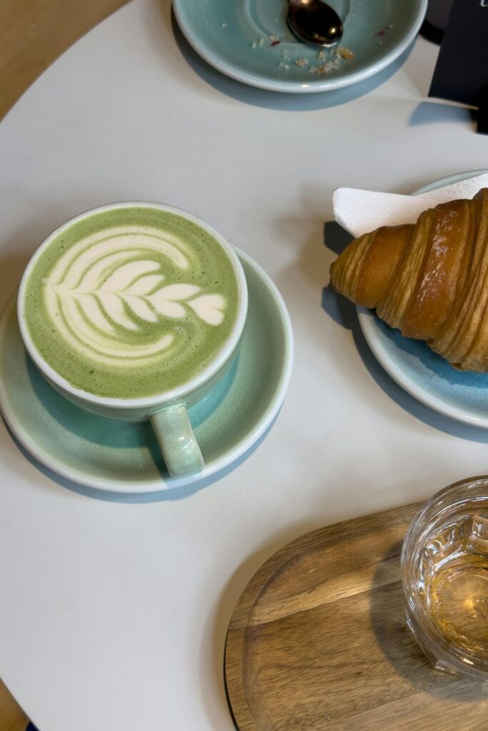 Felülről fotózott matcha latte csészében, mellette croissant és egy pohár ital az asztalon