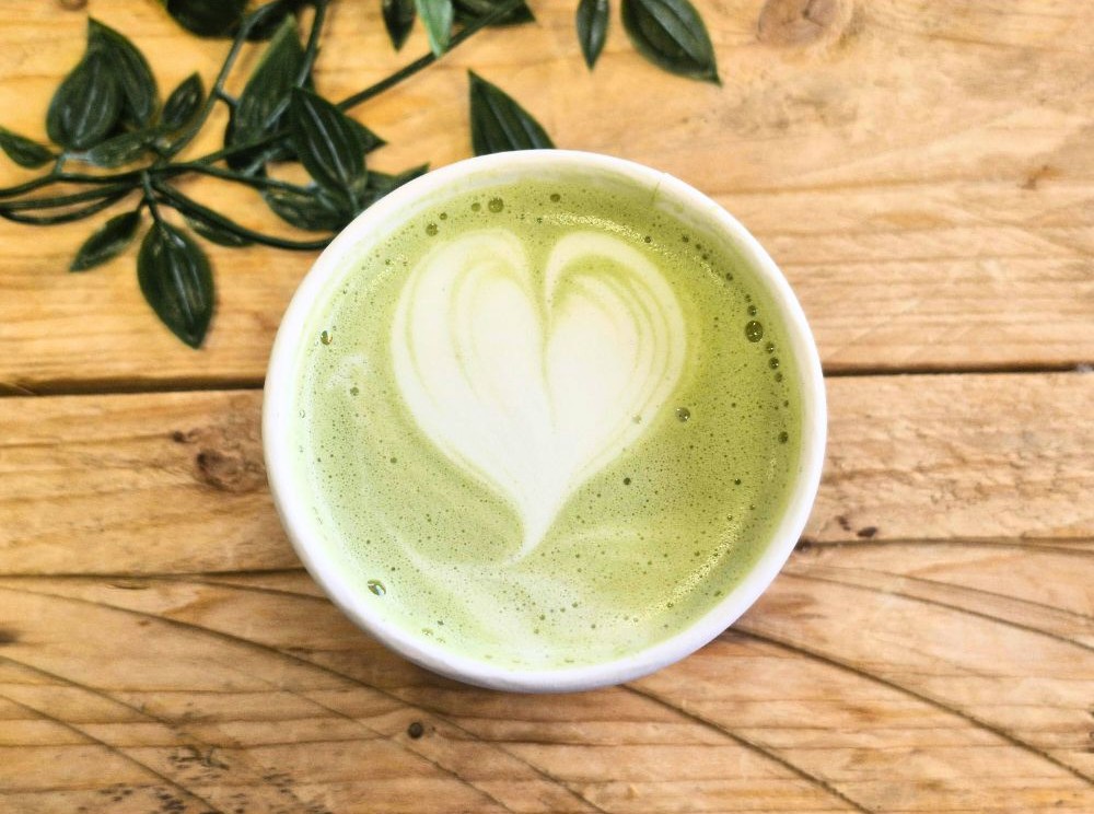 Matcha latte szív alakú tejhabbal felülnézetből, faasztalon, zöld levelek mellett