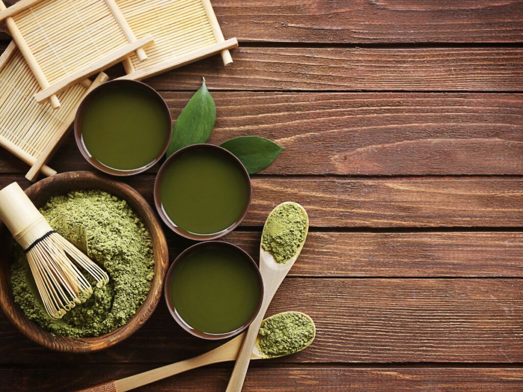 Három csésze elkészített matcha tea bambusz tálcán, mellette matcha por és bambusz habverő (chasen)