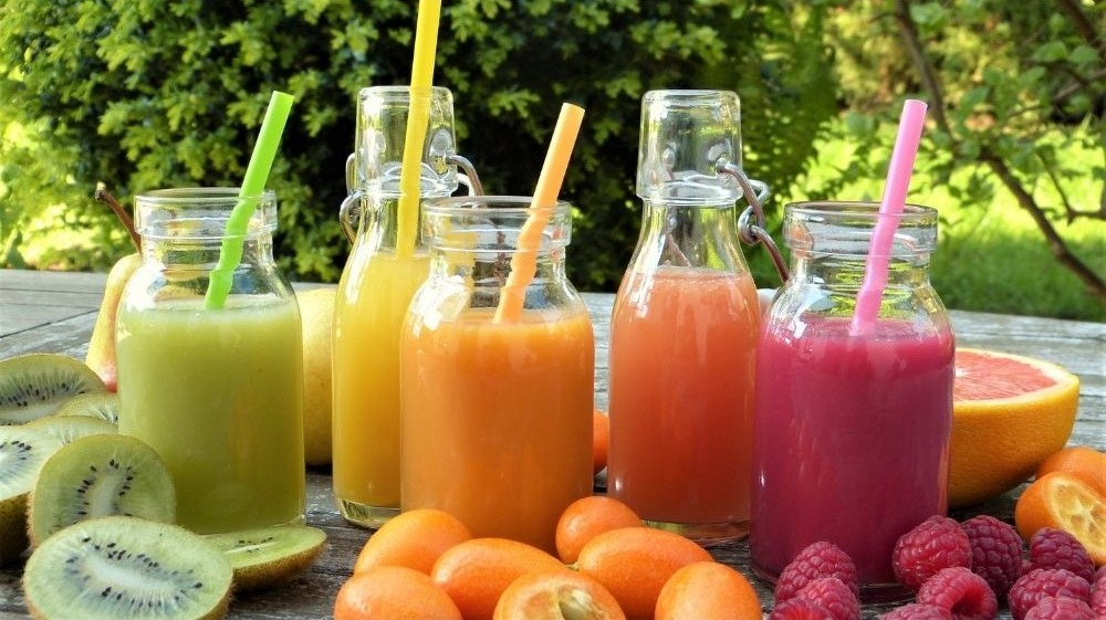 Öt különböző színű gyümölcs smoothie szívószállal, kiwivel, grapefruittal és málnával díszítve, kültéri asztalon